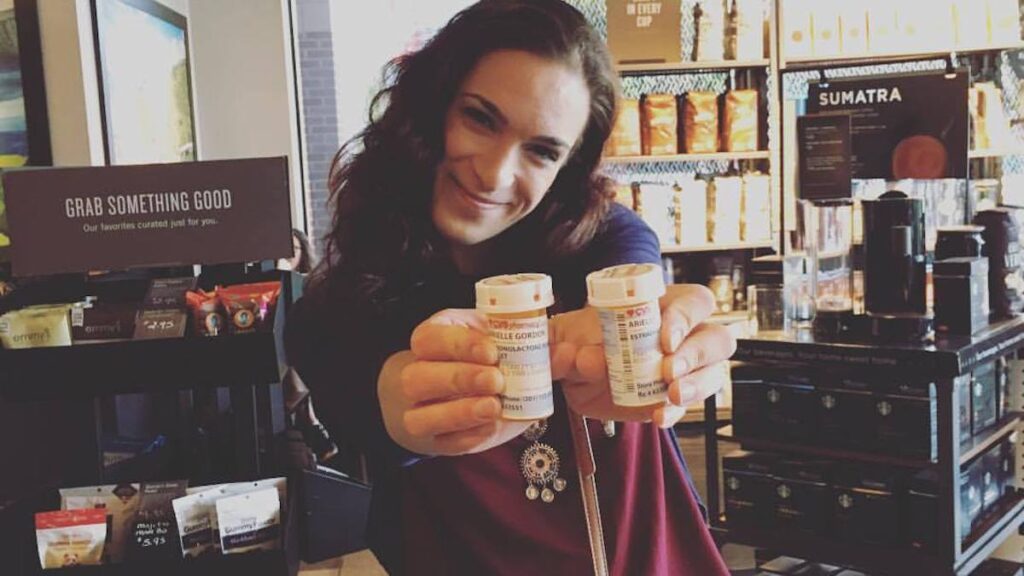 Arielle, a white trans woman with medium-length brown hair, holds two orange CVS pill bottles in front of her. She smiles and holds her body on an angle. She's inside of a Starbucks, standing in front of a wall stocked with bags of coffee.