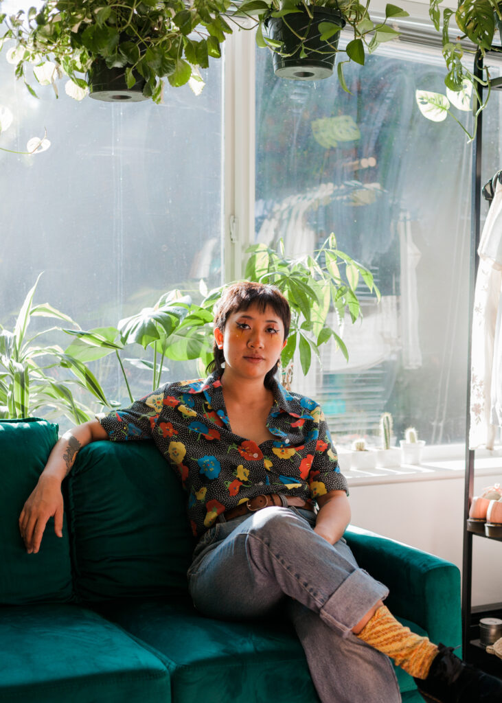 Felix sits on a turquoise couch, one arm posed across the back of the couch. He wears cuffed blue jeans and a polka-dot, floral print shirt. He has eyeliner applied around his eyes, forming the shape of a four-pointed star. Both behind and hanging above him are luscious green plants, including a number of succulents. 