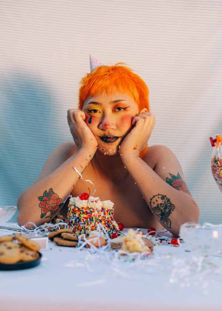 Felix has bright, orange hair and wears a polka-dot party hat. He is shirtless, with his shoulders visible above a white tablecloth. In front of him are numerous desserts, including a cake with rainbow sprinkles and a cherry, as well as chocolate chip cookies. His hands are pressed up against his chin. His makeup includes dark-colored lipstick, red blush, a teardrop drawn with black eyeliner, and winged eyeliner on his eyes.