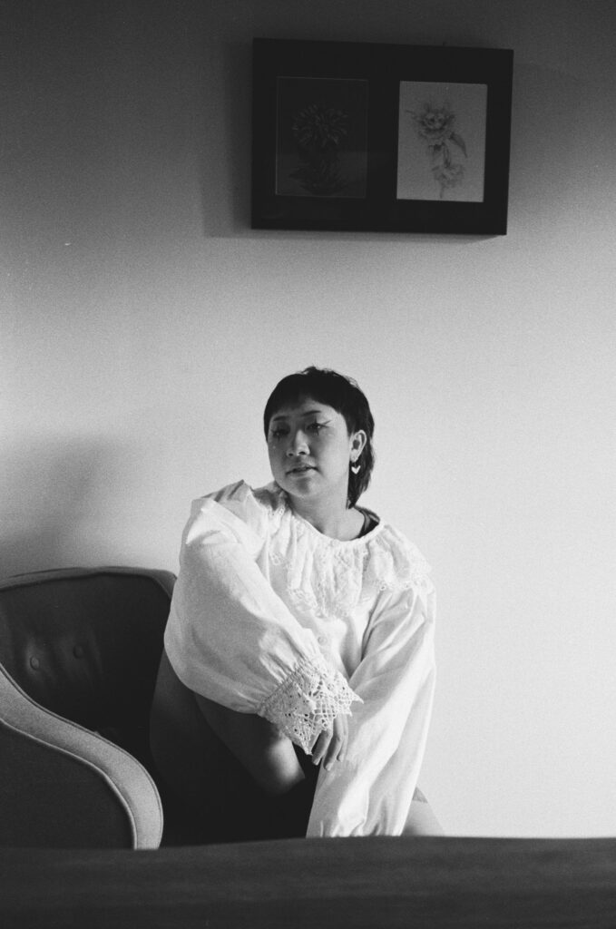 A grayscale image of Felix wearing a white blouse, sitting in an armchair. He holds his head on an angle, his eyes turned toward the camera. He has black eyeliner, drawn to resemble a four-pointed star.