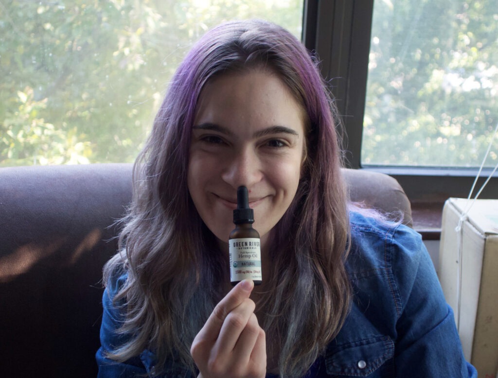Arielle, a white trans woman with purple hair, displaying a bottle of CBD oil she used while recovering from bottom surgery.
