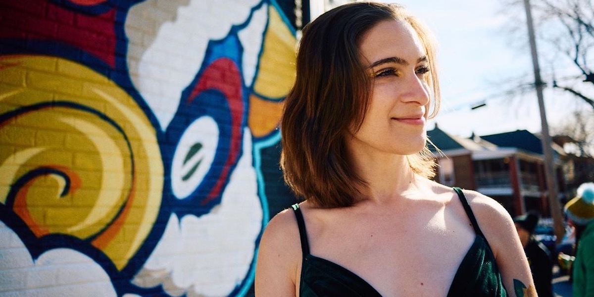 Arielle, a white nonbinary person with shoulder-length wavy brown hair, wearing a green velvet v-neck dress and looking off to the side while standing in front of a colorful abstract mural.