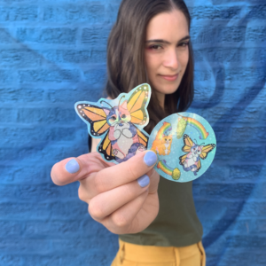 A white nonbinary person wearing yellow pants and a green tanktop stands in front of a blue brick wall. They're holding two stickers: one a cat-butterfly hybrid with orange and yellow triangle-patterned wings with a black outline. The cat body has pink, purple, and blue stripes with a white belly, paws held to their chest, and a stripes tail held over their feet. The other the lifecycle of a cat-erpillar.