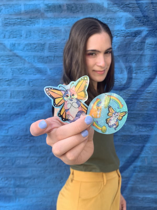 A white nonbinary person wearing yellow pants and a green tanktop stands in front of a blue brick wall. They're holding two stickers: one a cat-butterfly hybrid with orange and yellow triangle-patterned wings with a black outline. The cat body has pink, purple, and blue stripes with a white belly, paws held to their chest, and a stripes tail held over their feet. The other the lifecycle of a cat-erpillar.