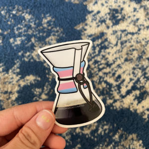 A white hand holds Chemex coffeemaker sticker with the transgender pride flag superimposed over the wooden handhold. A blue carpet is below.