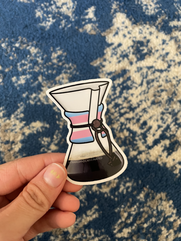 A white hand holds Chemex coffeemaker sticker with the transgender pride flag superimposed over the wooden handhold. A blue carpet is below.